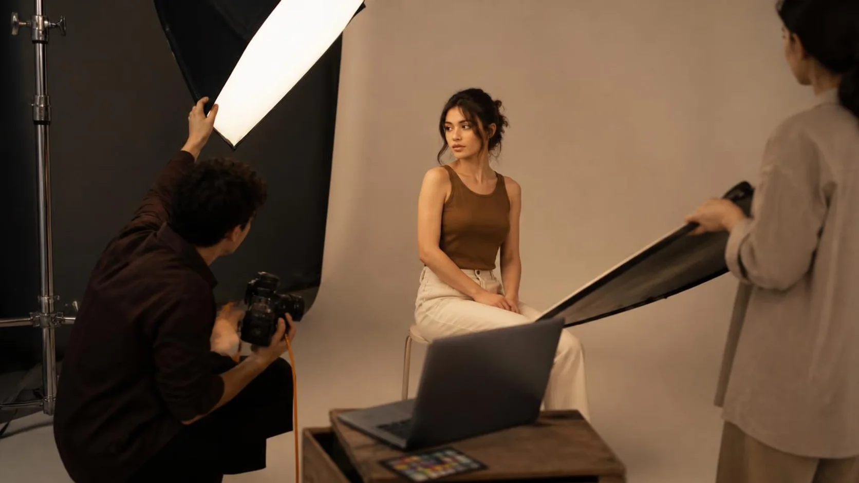 Fotógrafa ajusta softbox durante ensaio de retrato em estúdio, com modelo sentado diante do fundo neutro.